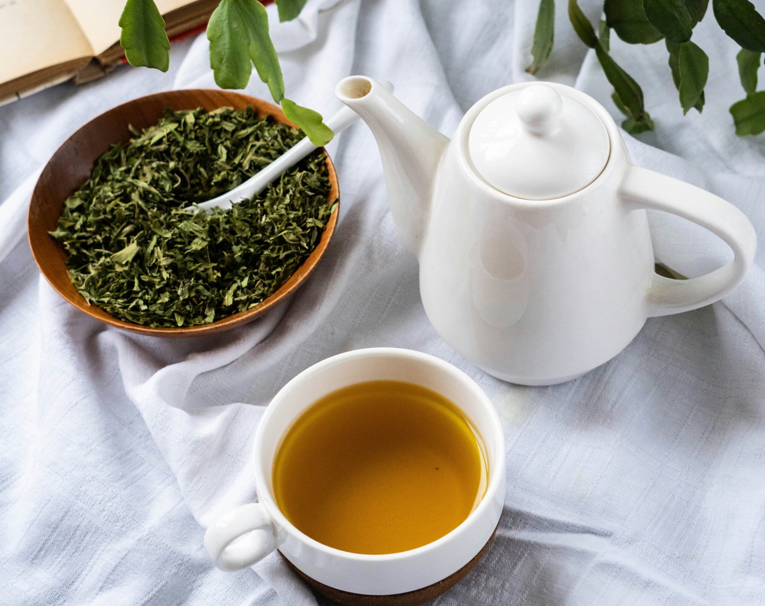 Uma xícara de chá verde servida sobre uma toalha branca ao lado de uma tigela com folhas de chá verde amassadas e uma chaleira branca.